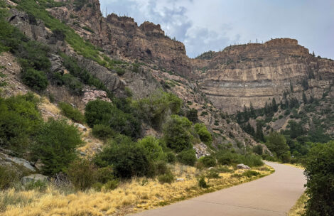 A paved bike path winds through Glenwood Canyon, which is lined with dry grasses and green shrubs beneath steep cliffs and an overcast sky.
