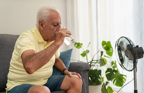 Senior man refreshing himself drinking water and using an electric fan during a hot day at home.