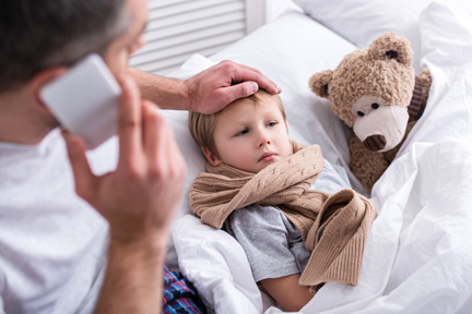 Sick child in bed with his Teddy bear and a concerned father on the phone checking his temperature with his hand.