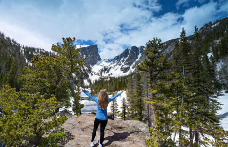 Joyful girl with raised hands relaxing on top of the mountain, looking at beautiful summer landscape.