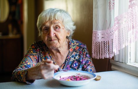 An elderly woman eating soup sitting at a table in the house.