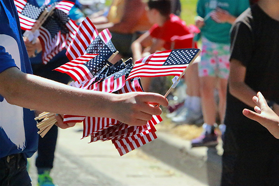 A person handing out American flags along parade route, hand reaching from corner of frame, blurred crowd in background. Independence Day celebration, 4th of July in small town USA.